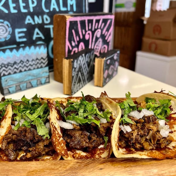 Three Folklore Artisanal Mexican tacos with meat, cilantro, and onions on a wooden board.