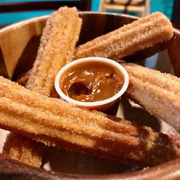 A wooden bowl filled with golden-brown churros dusted with sugar, alongside a cup of caramel dipping sauce.