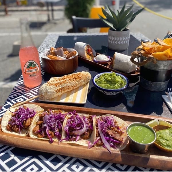 Folklore Artisanal Mexican spread with tacos, burritos, elote, guacamole, chips, churros, and a Jarritos.