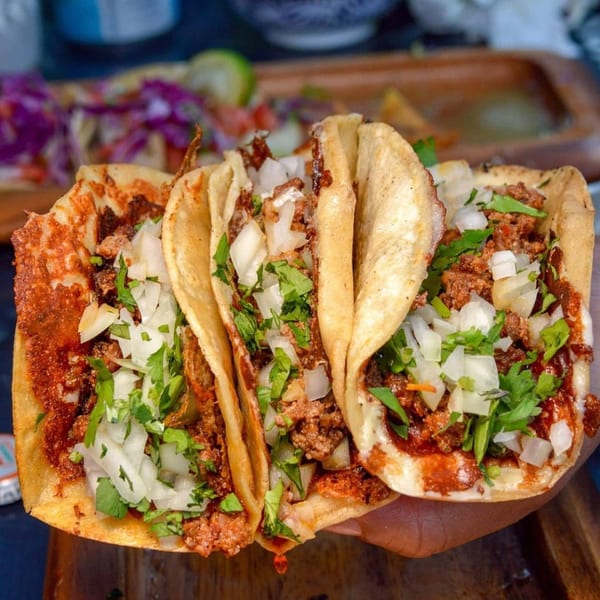 Three Folklore Artisanal Mexican tacos with meat, onions, and cilantro, held over a wooden tray.