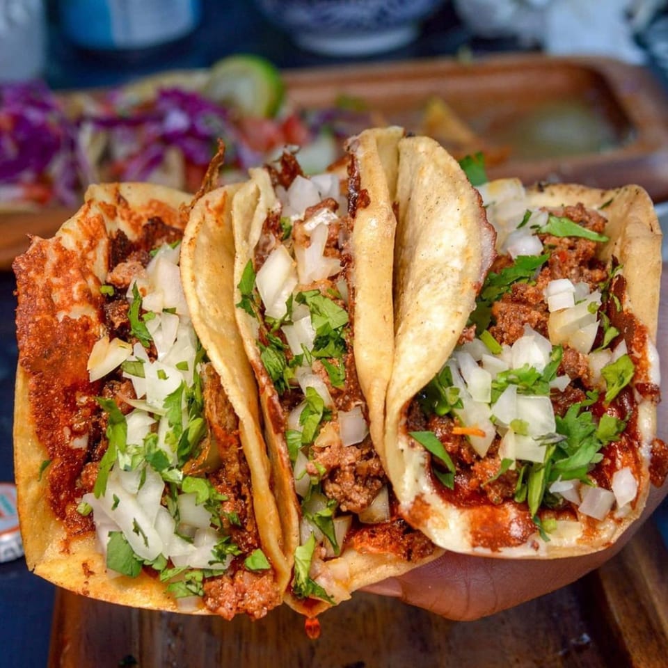 Three Folklore Artisanal Mexican tacos with meat, onions, and cilantro, held over a wooden tray.