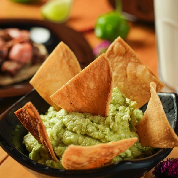 A black bowl of guacamole with several tortilla chips standing upright in it, on a wooden table.